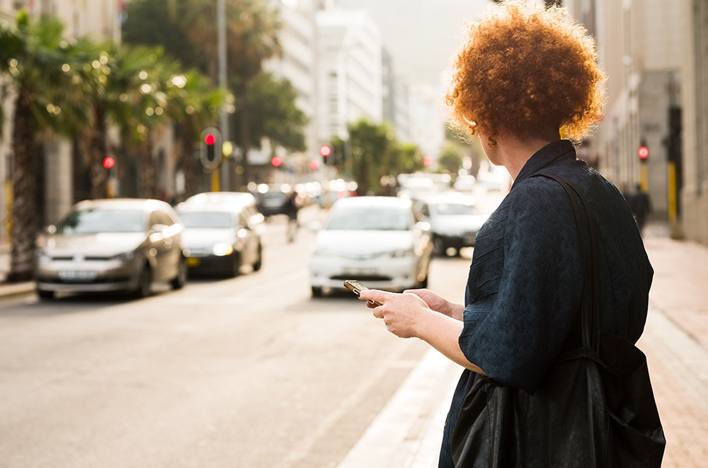 一个成熟的女人站在城市的街道上，拿着手机等出租车＂itemprop=
