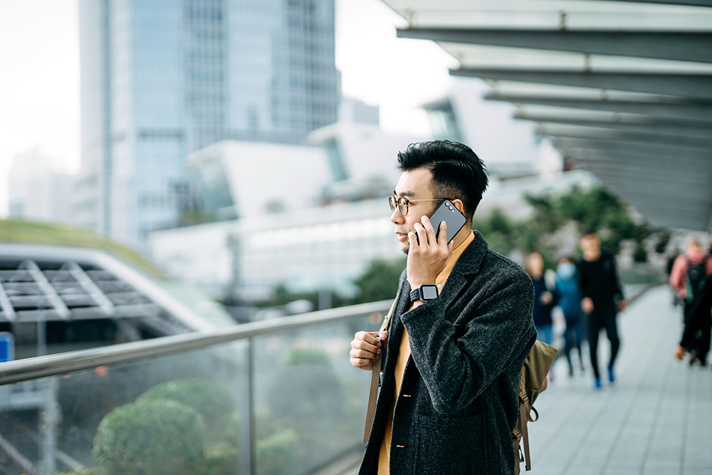 年轻的亚洲城市商人在城市通勤时用智能手机通话，单肩背着背包