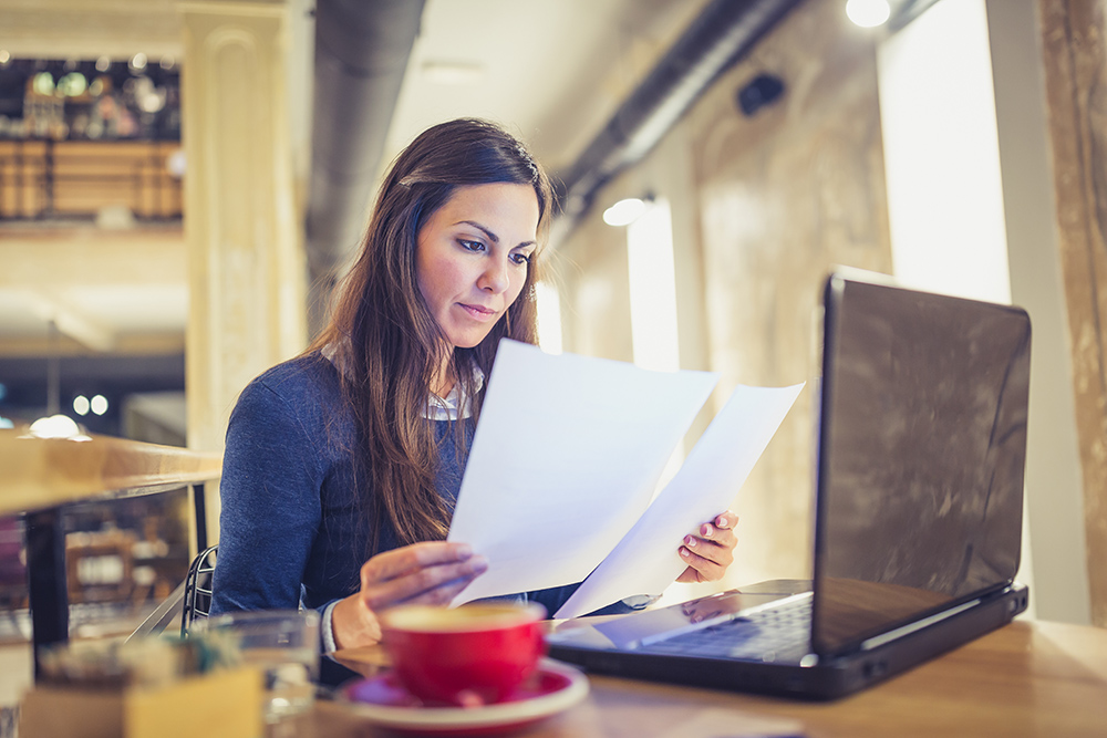 漂亮的黑发年轻女子坐在咖啡馆，喝咖啡，浏览文件，使用笔记本电脑