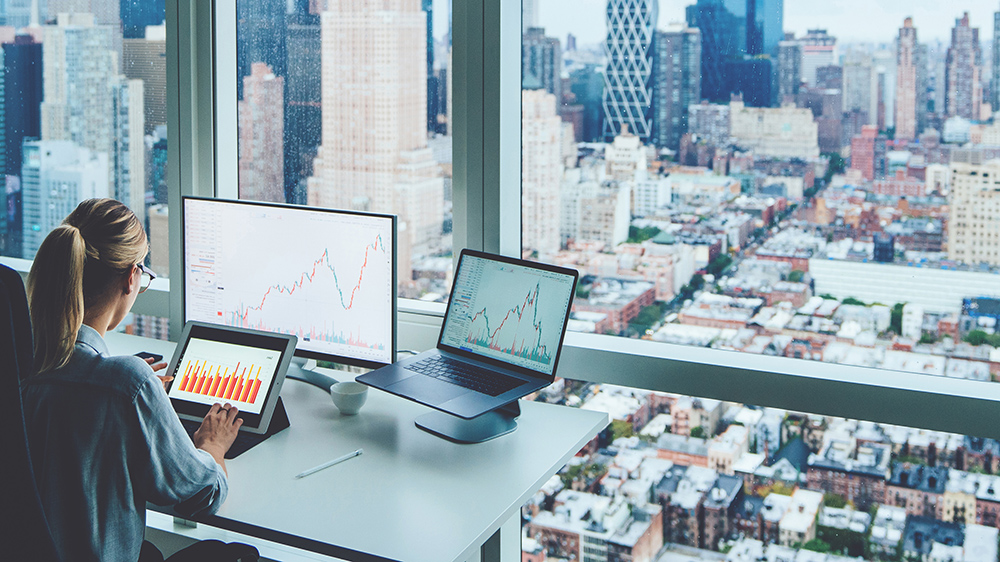 商业妇女坐在全景摩天大楼办公室桌面前PC电脑与财务图表和统计数据的显示器的背影。数字市场分析与区块链投资